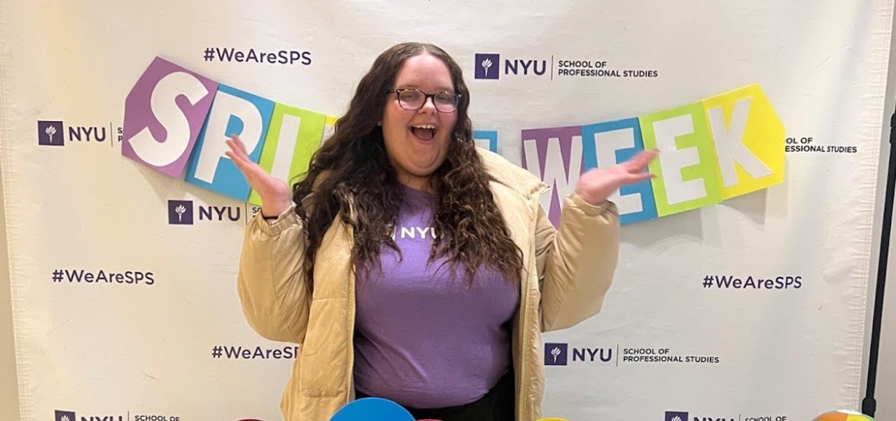 Payten Slack, student at the Jonathan M. Tisch Center of Hospitality, NYU SPS, poses for a photo in front of a Spirit Week step-and-repeat covered in NYU logos.