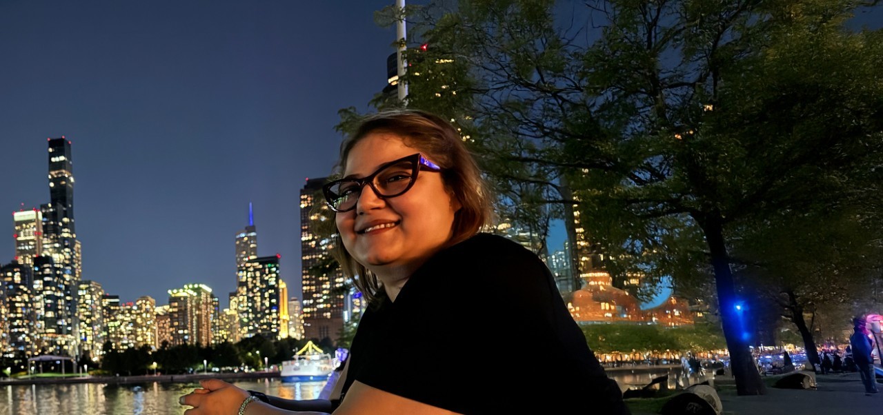 Reem Okar, student at the Center for Publishing and Applied Liberal Arts, NYU SPS smiles for the camera while dining at an outdoor restaurant with a view of the Manhattan skyline in the background.