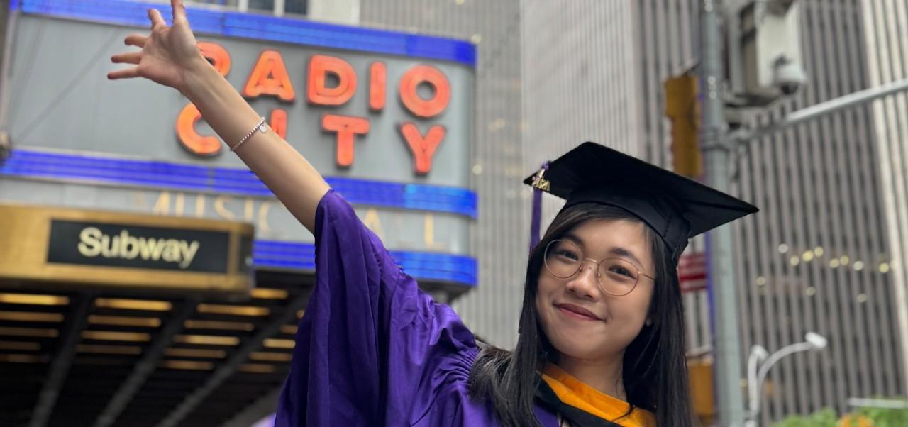 Ruby Chan, student at the Center for Publishing and Applied Liberal Arts, NYU SPS, poses for a photo in front of Radio City Music Hall while dressed in her cap and gown for graduation.