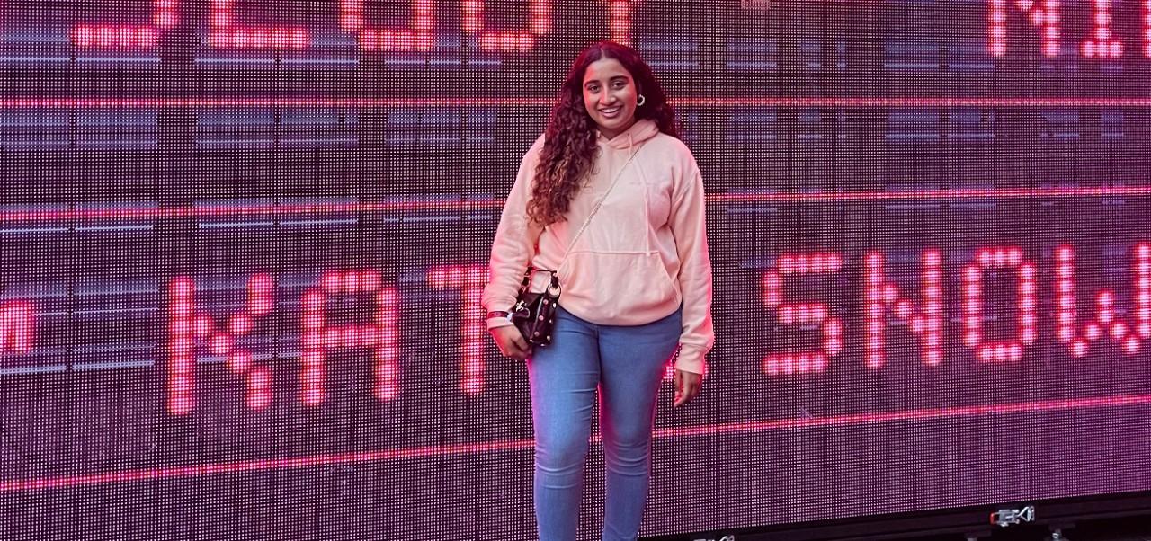 Stuti Daga, student at the Robert Tisch Institute for Global Sport at NYU SPS, smiles for the camera while standing in front of a massive digital display conatining pink letters.