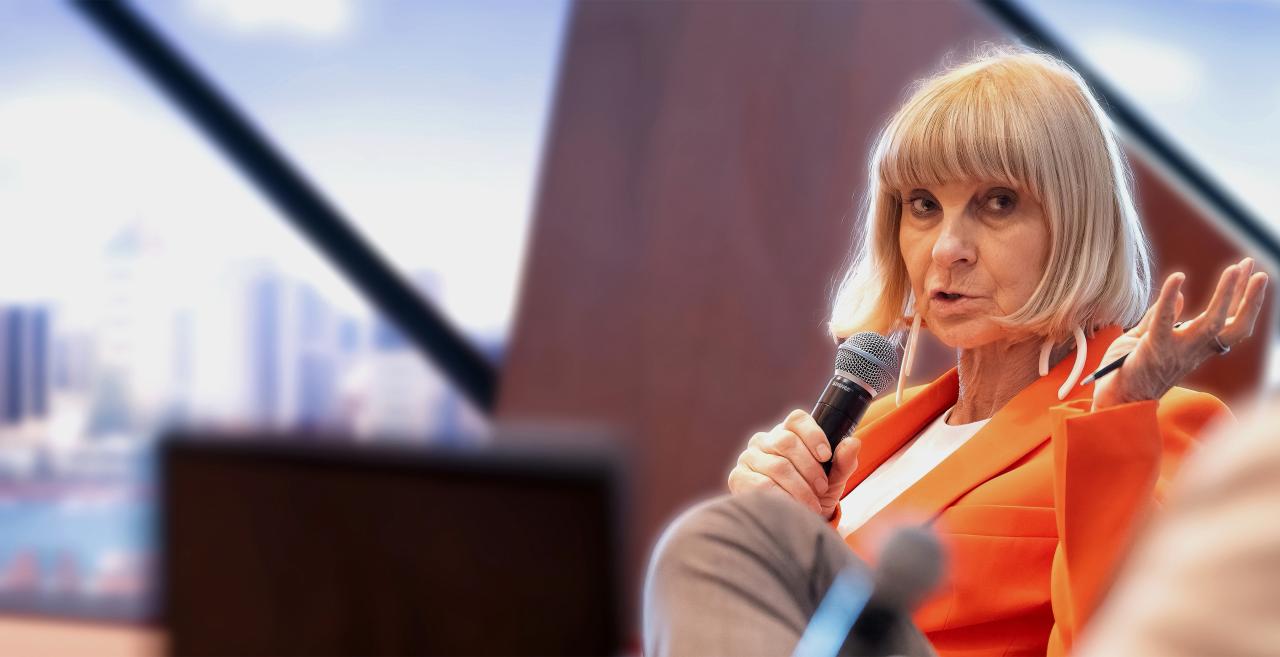 Anna Tavis speaking into a microphone during a panel discussion with a city skyline in the background