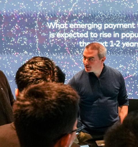 An inquisitive gentleman addresses a room of students while giving presentation in front of a brighly lit Powerpoint slide.
