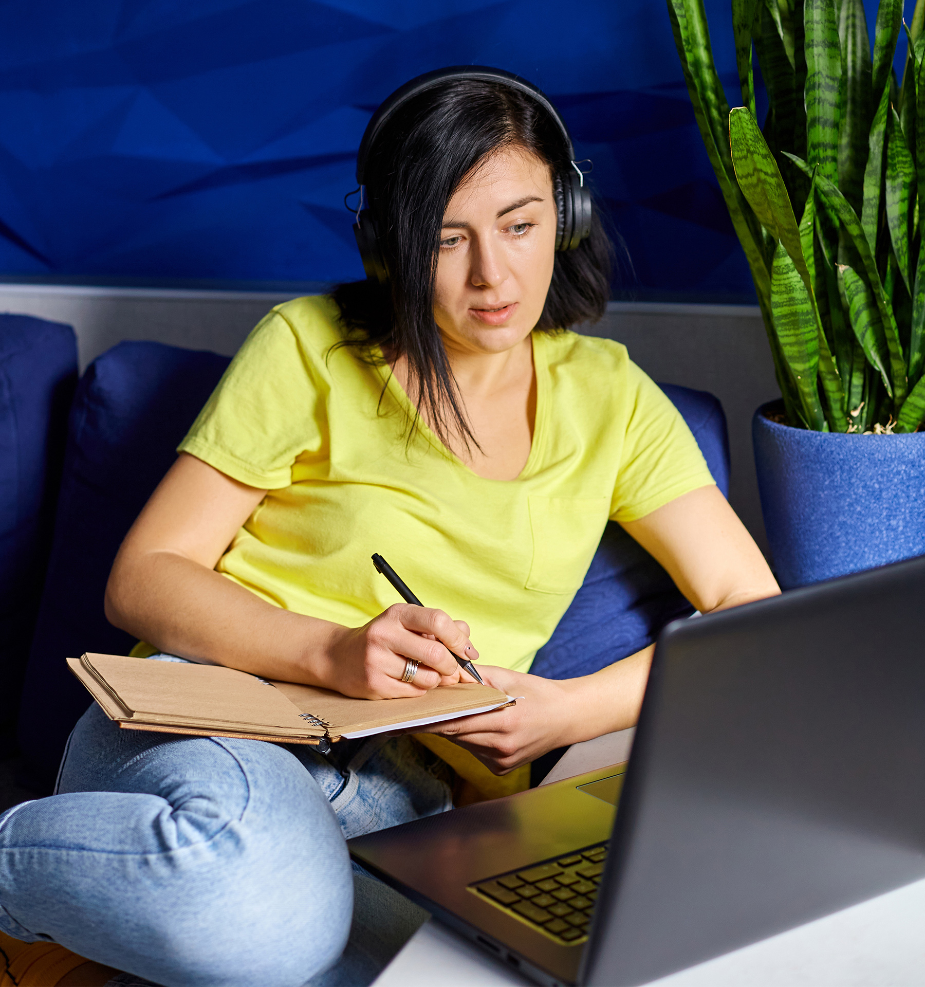 Student sitting on a blue couch with a notebook, pen, and headphones, taking notes in front of an open laptop.