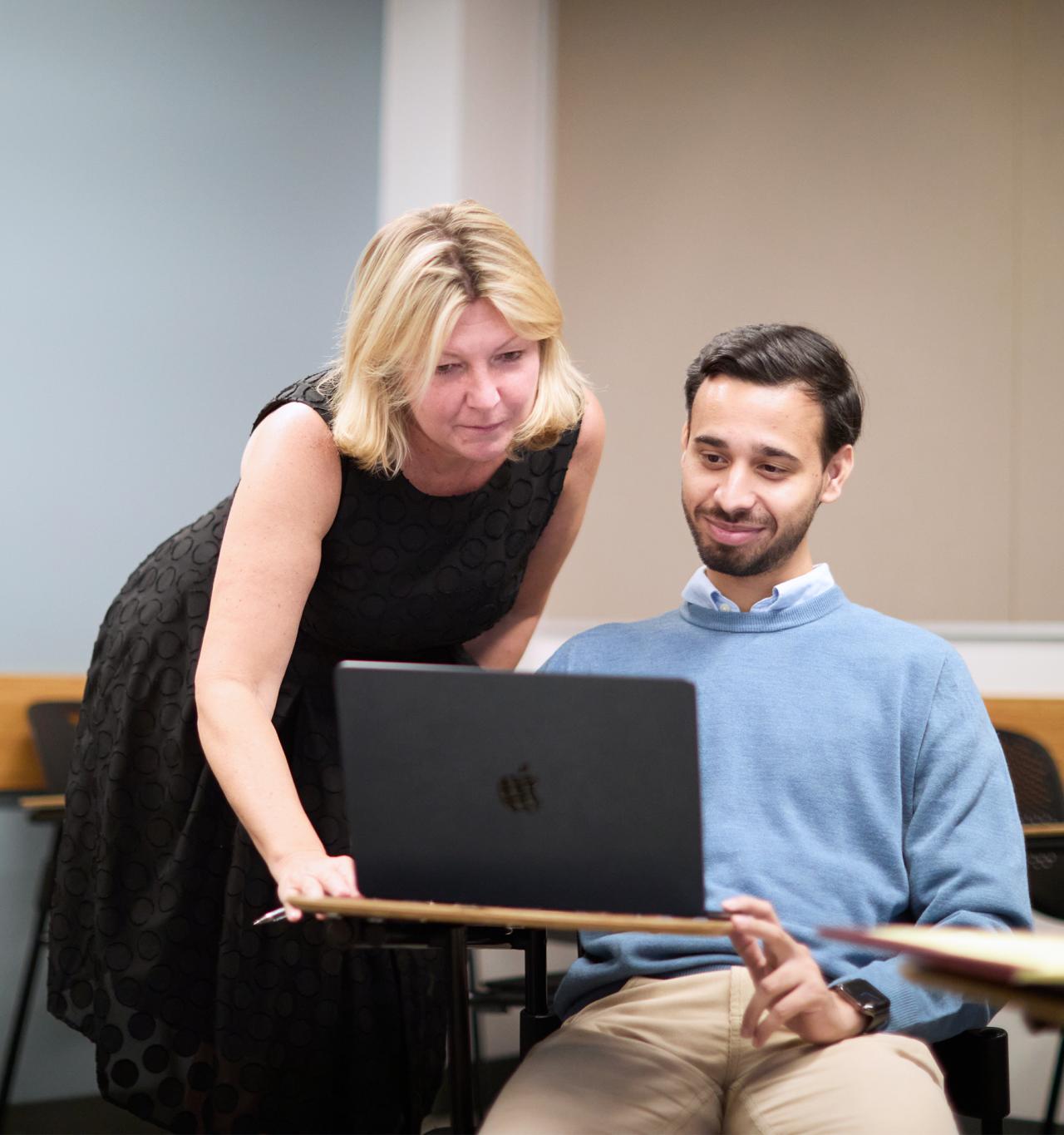 A faculty mentor in the NYU SPS mentoring program helps a student who sits at a desk with a laptop