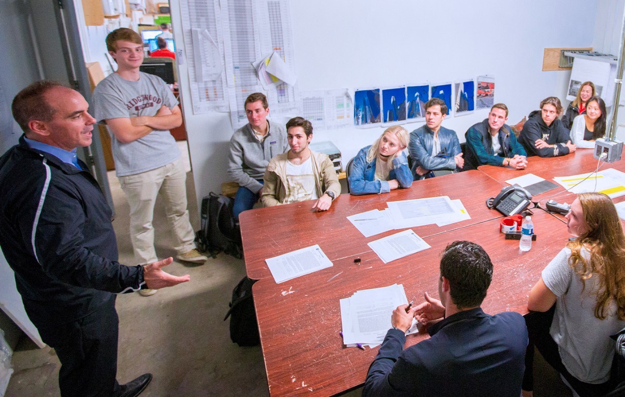 A group of people sit around a large table in a meeting room, attentively listening to a man speaking at the end of the table.