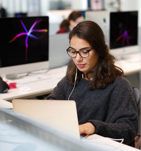 A woman with glasses, wearing earphones, focuses on her laptop in a modern office filled with several iMac computers.