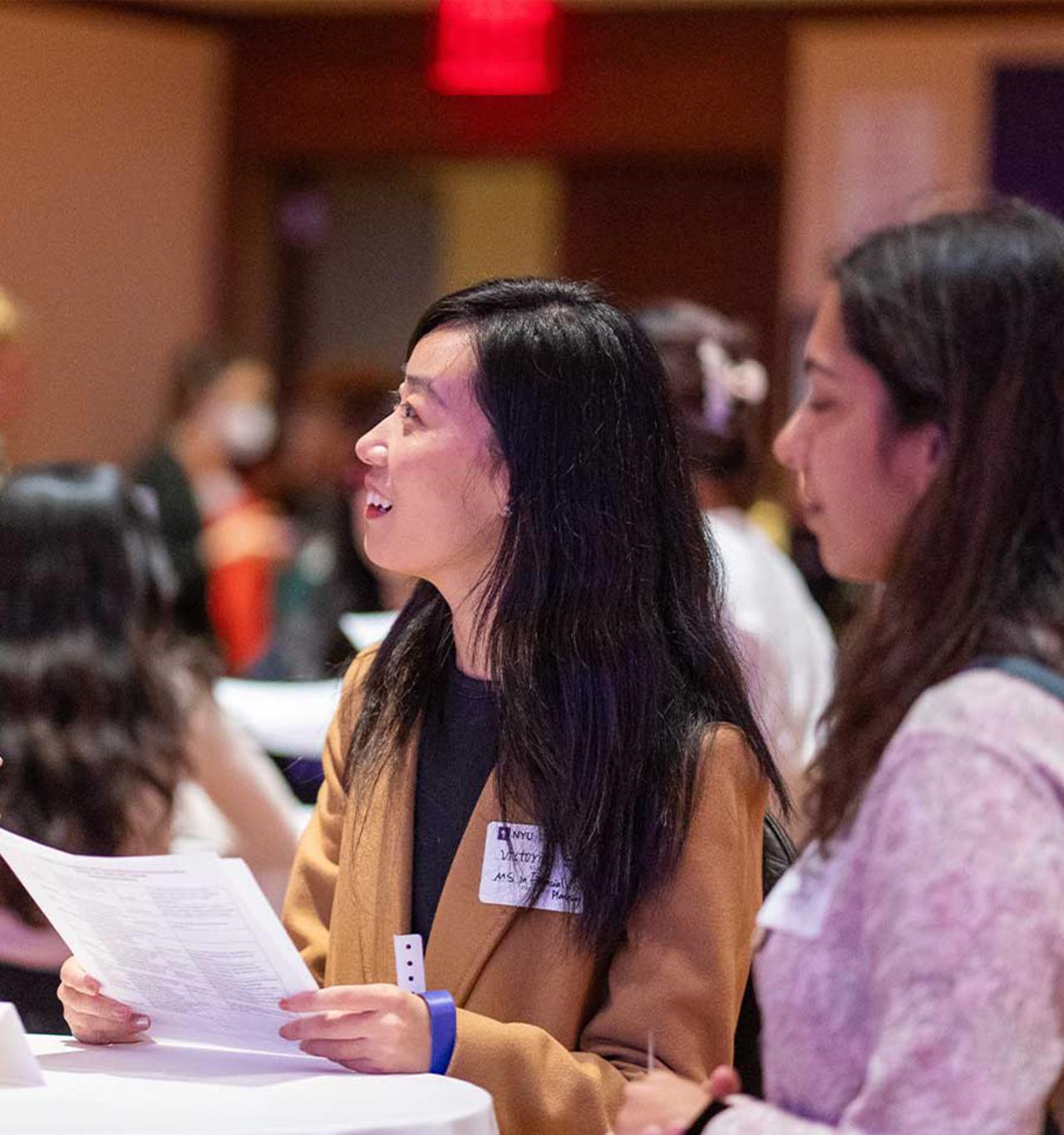 Three women engage in conversation at a networking event, around a tall table labeled "1." 