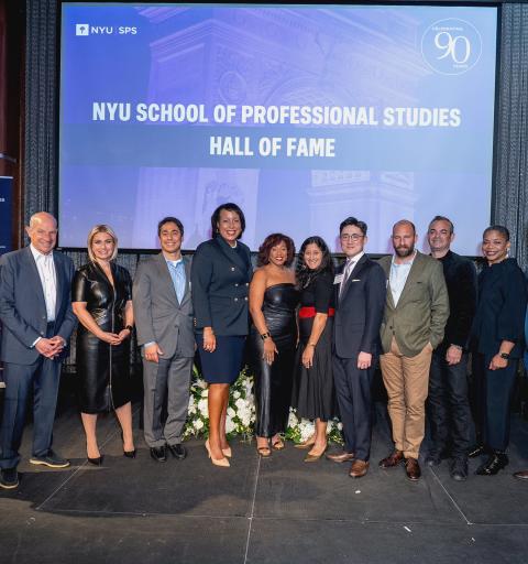Group of people standing on stage at the NYU School of Professional Studies Hall of Fame event, with banners, floral arrangements, and a backdrop reading “Celebrating 90 Years.”
