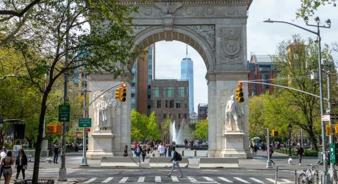Historic arch in a park with sculptures, surrounded by green trees and urban buildings. A fountain is visible in the background; people walk nearby.