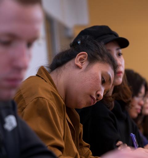 A female NYU SPS hospitality scholarship student leans her head to the side while taking notes during a lecture with other students on either side of her