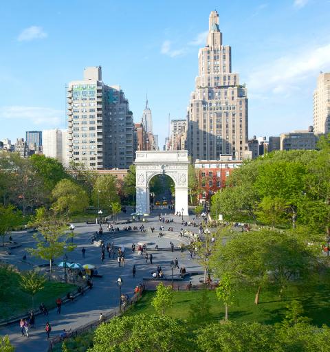 An aerial daytime shot of Washington Square Park facing north, with the 5th avenue corridor extending beyond the iconic white arch in the distance and pedestrians milling about in the central plaza.