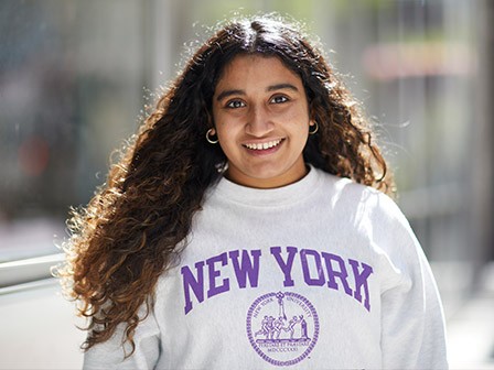 Smiling NYU student wearing a white “New York” sweatshirt, standing outdoors on campus.