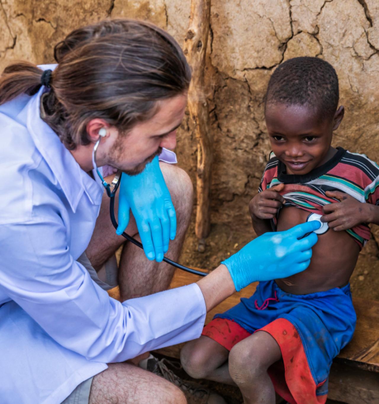 A doctor wearing blue gloves and a white coat examines a smiling child with a stethoscope in a rural setting.