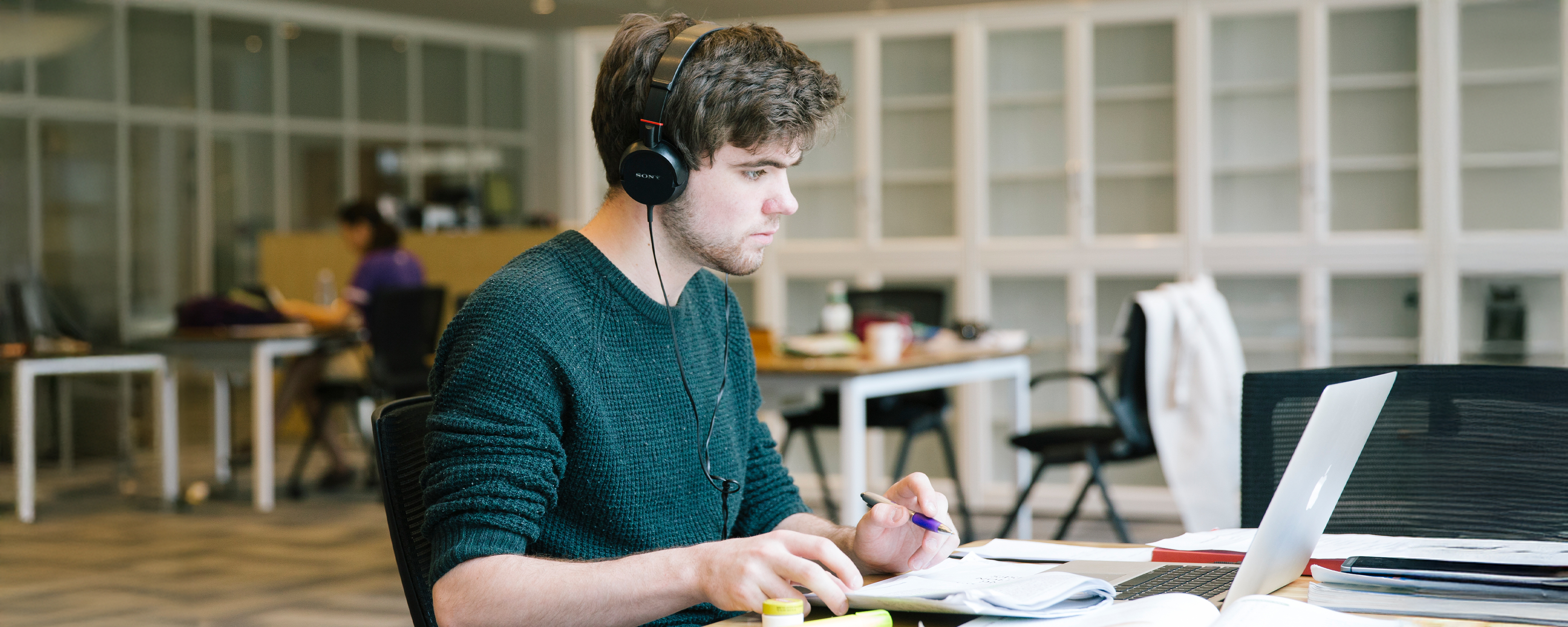 A focused young man in headphones studies at a table with a laptop, open books, and a highlighter.