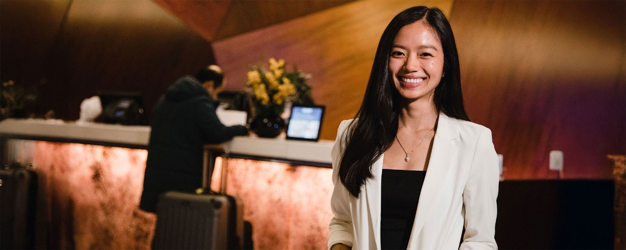 Woman smiles in front of the front desk of a hotel lobby to represent AAS in Hospitality's Your Experience and Our Experience