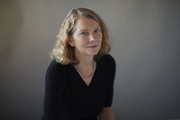 Front profile of Jenny Mcphee: Female presenting person with blue eyes and blond hair wearing a necklace of pearls and a black dress in front of a gray background.