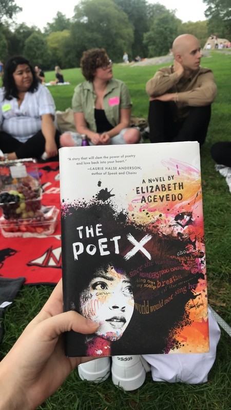 The book cover of The Poet X by Elizabeth Acevedo is shown in the forefront with 3 seated students in the background.