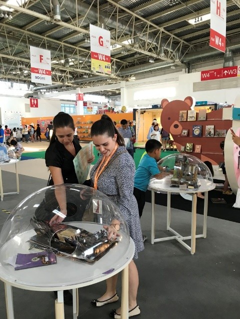 2 students engaged with a 3D popup book, inside a plastic dome, with holes to put their arms inside of