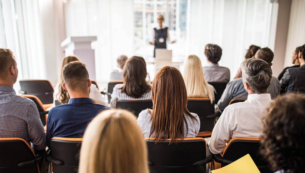 A rear view of master’s degree students sitting in chairs in a seminar room while watching and listening to a person deliver a speech.