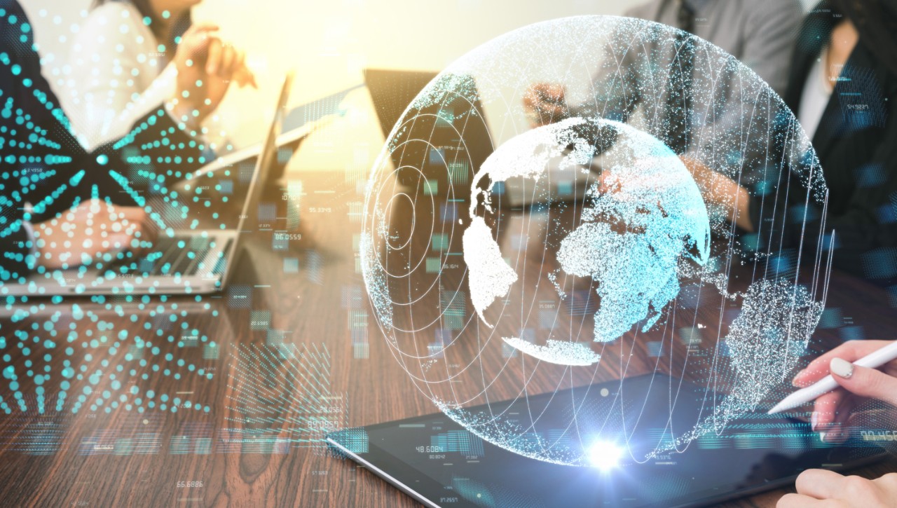 A futuristic holographic vision and global network appears above a tablet on a table during a meeting with multiple people.