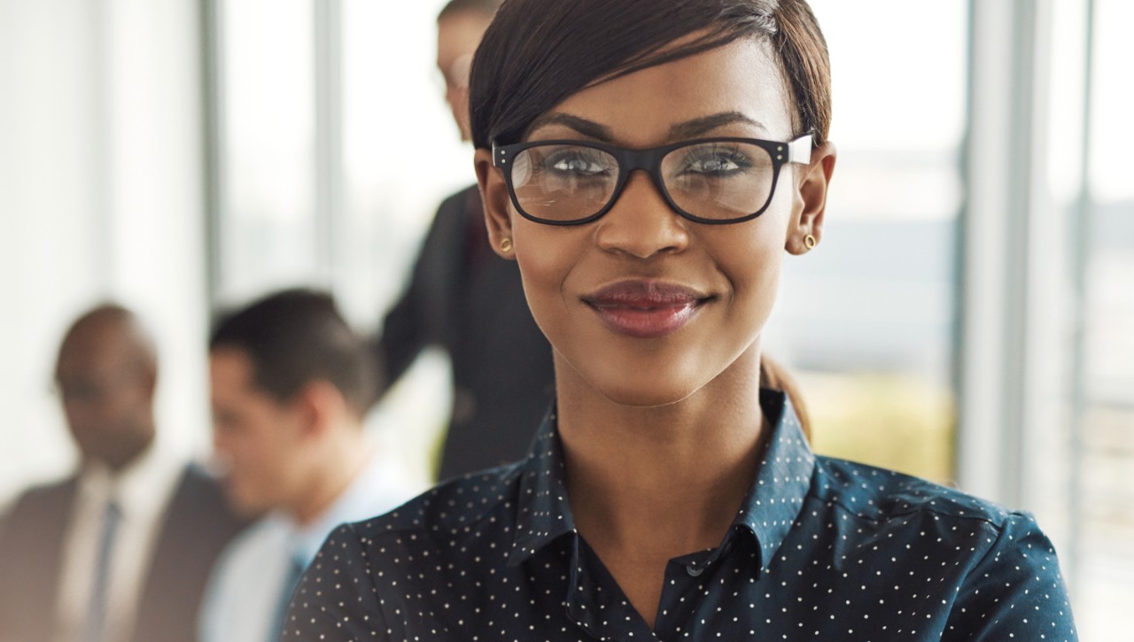 A closeup of a woman executive with glasses in the foreground and other workers blurred out in the background.