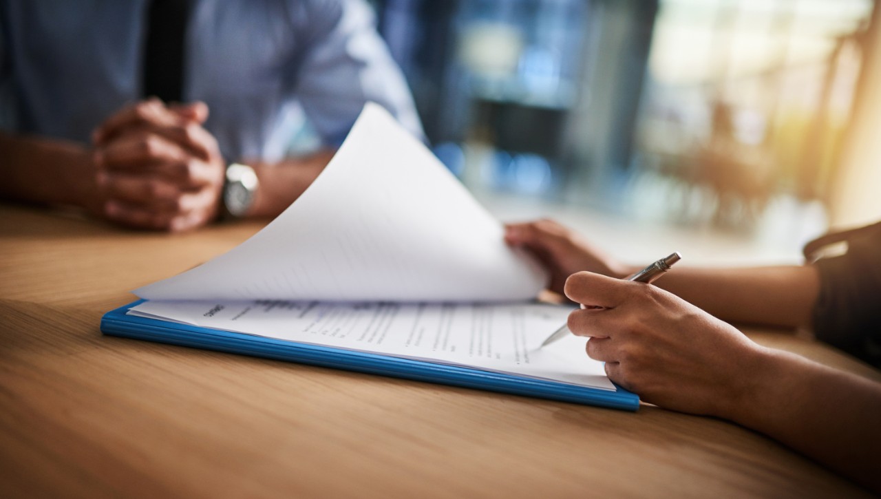 A left-handed professional writer points a pen on a page in a packet during a meeting with another person at the table.