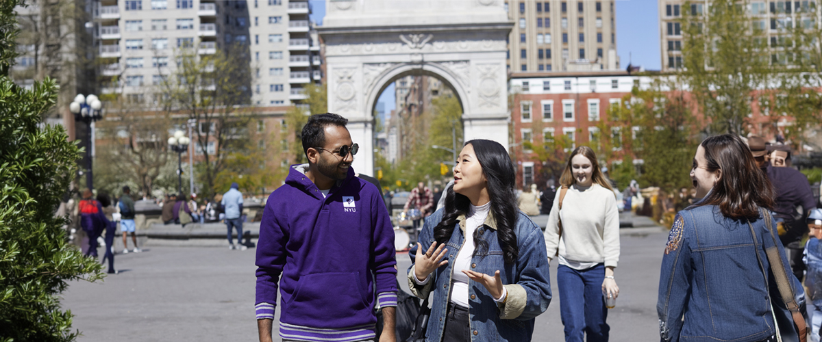 Two NYU SPS students, one male wearing a purple NYU hoodie and one female wearing a jean jacket, have a conversation while walking through a busy Central Park in NYC