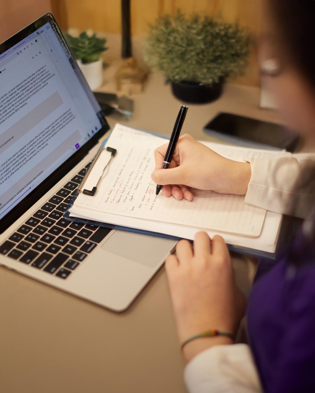 A person exploring online undergraduate degrees at NYU SPS takes notes on a sheet of paper in front of a laptop