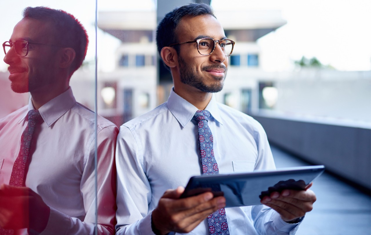 A man leans against a window with a tablet in his hands while looking into the distance